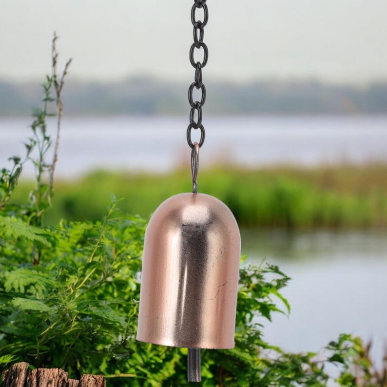 May include: A copper-colored bell hanging from a black chain. The bell has a rounded top and a flared bottom. The background features green foliage and a body of water, creating a natural setting.