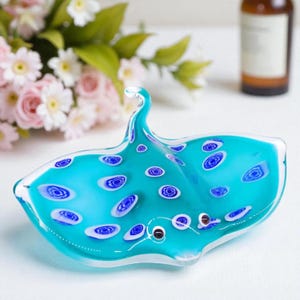 Może przedstawiać: Turkusowa szklana dekoracyjna miska w kształcie płaszczki. Miska ma białe i niebieskie okrągłe wzory na powierzchni. Płaszczka ma zakrzywiony ogon i małe czarne detale oczu.
