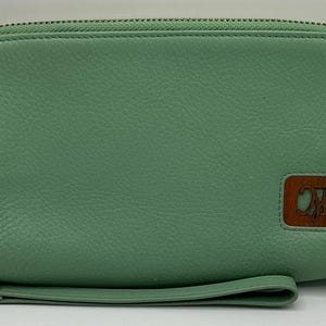 Op de afbeelding: Een groene leren clutch met rits en een houten logo met de tekst "CM". De clutch heeft een polsbandje.