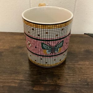 May include: A ceramic mug with a butterfly design. The mug features a pink band with a butterfly and flower pattern, set against a grid of white and black squares. The top and bottom sections have a gold and black grid pattern.
