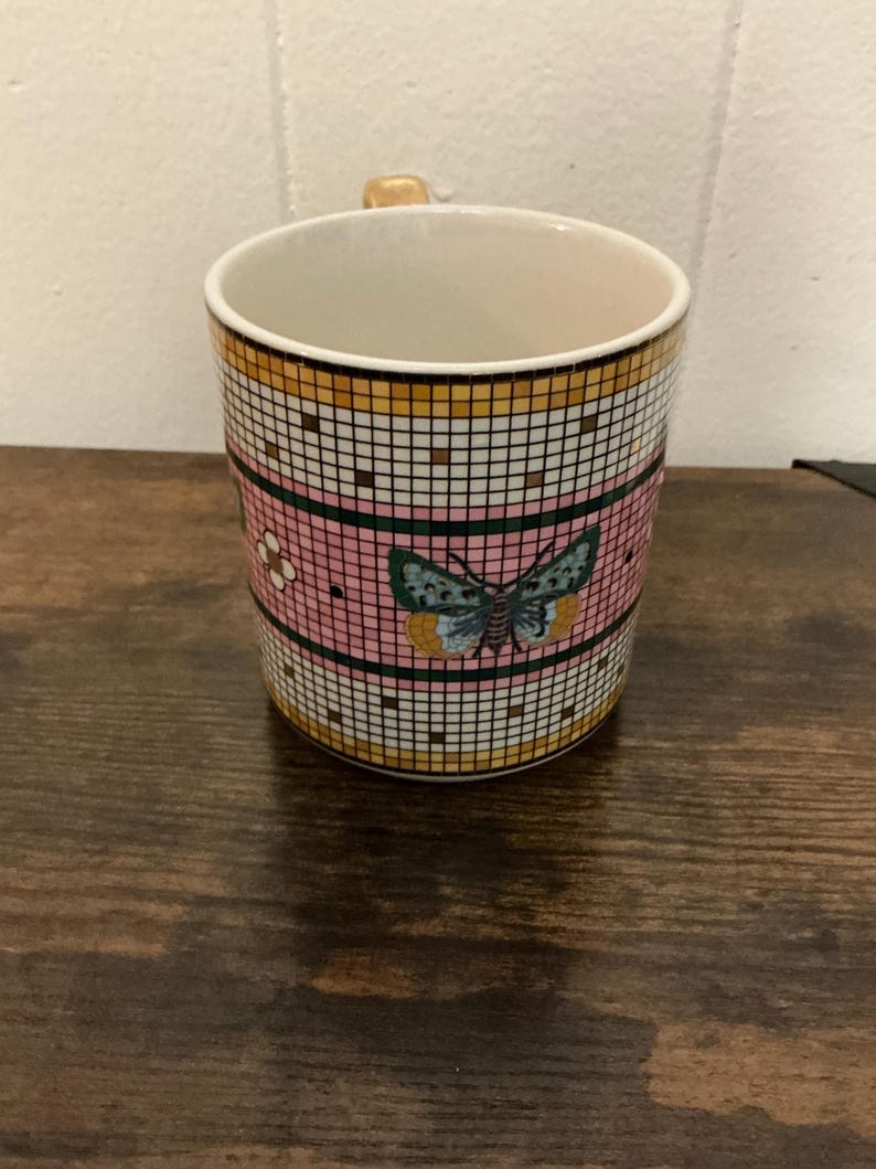 May include: A ceramic mug with a butterfly design. The mug features a pink band with a butterfly and flower pattern, set against a grid of white and black squares. The top and bottom sections have a gold and black grid pattern.