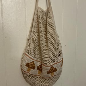 May include: A cream-colored cotton string market bag featuring a wide, solid-knit horizontal band near the bottom. This band is decorated with a repeating pattern of brown and tan mushrooms, and the bag includes two long shoulder straps.