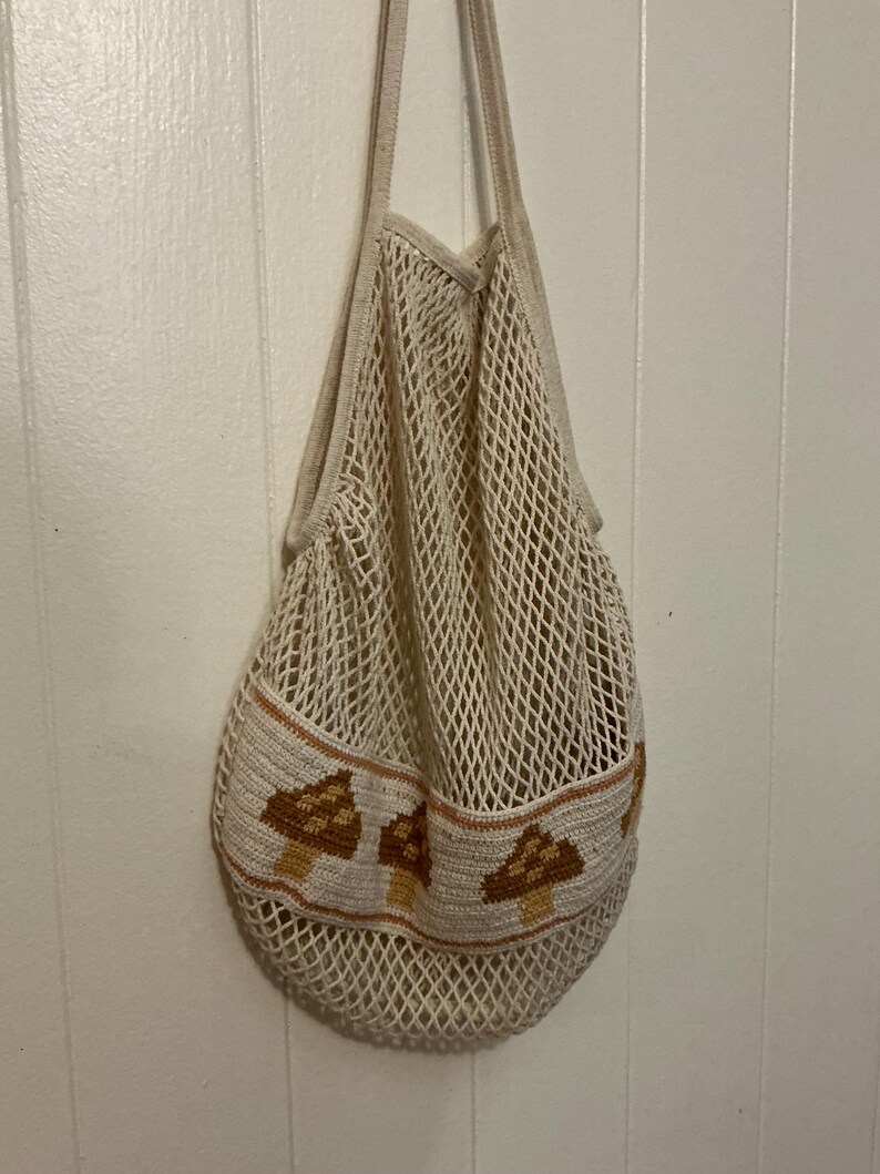 May include: A cream-colored cotton string market bag featuring a wide, solid-knit horizontal band near the bottom. This band is decorated with a repeating pattern of brown and tan mushrooms, and the bag includes two long shoulder straps.