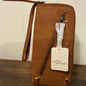 May include: A brown leather wallet with a wrist strap and a zipper closure. The wallet has a tag that says "free people" and "www.freepeople.com". The wallet is rectangular and has gold-colored metal accents.