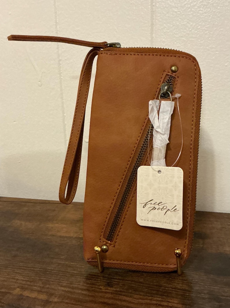 May include: A brown leather wallet with a wrist strap and a zipper closure. The wallet has a tag that says "free people" and "www.freepeople.com". The wallet is rectangular and has gold-colored metal accents.