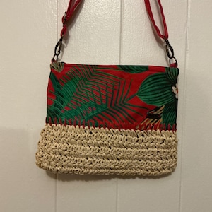 May include: A red and green tropical print crossbody bag with a woven raffia bottom. The bag has a red strap and bronze-colored hardware. The bag is hanging against a white background.