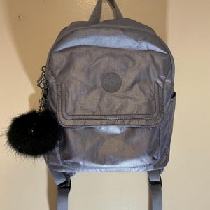 May include: A silver-gray backpack with a small front pocket and a black fluffy pom-pom attached. The backpack has adjustable straps and a top handle. A circular logo is centered on the front.