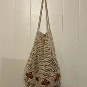 May include: A beige cotton string market bag featuring a solid woven band at the base decorated with a repeating pattern of brown and tan mushrooms. The bag has an open mesh net construction and two long shoulder straps for carrying items.