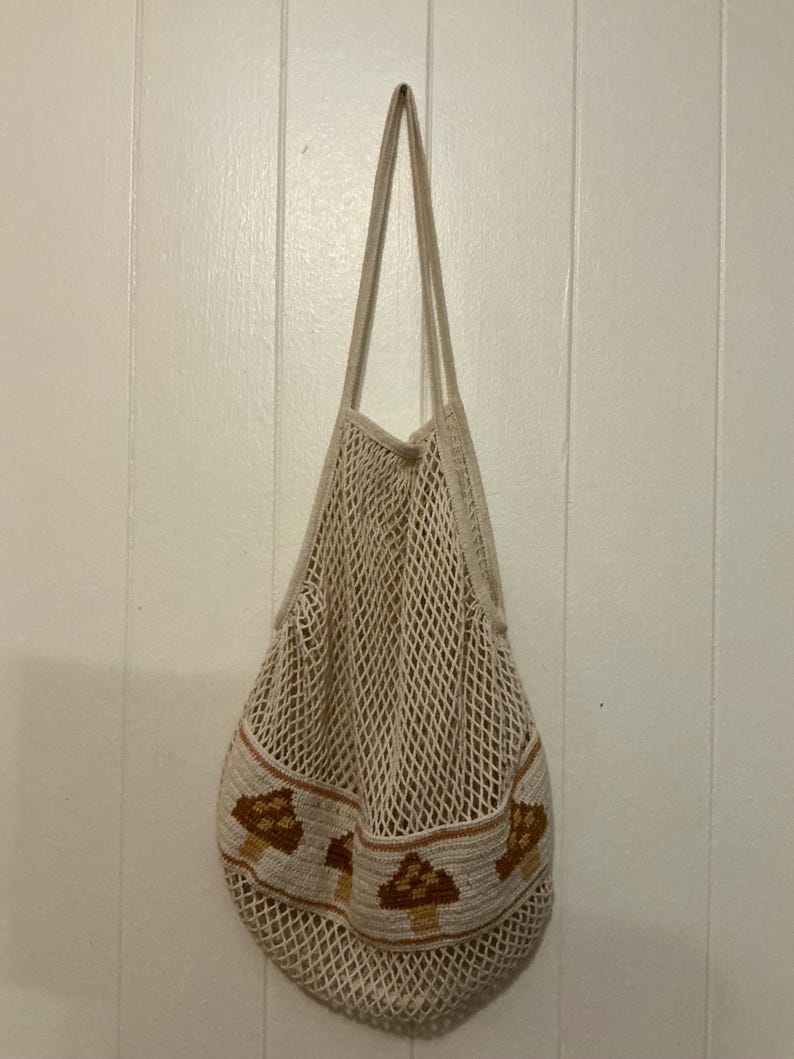 May include: A beige cotton string market bag featuring a solid woven band at the base decorated with a repeating pattern of brown and tan mushrooms. The bag has an open mesh net construction and two long shoulder straps for carrying items.
