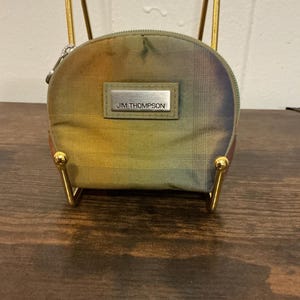Puede incluir: Un peque&ntilde;o monedero redondo con cierre de cremallera. El bolso presenta un estampado de cuadros en tonos verdes, amarillos y naranjas. Una etiqueta rectangular plateada con el texto "JIM THOMPSON" est&aacute; centrada en la parte delantera. El bolso se exhibe sobre un soporte met&aacute;lico dorado.