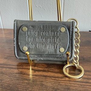 May include: A dark grey wallet with a gold chain and key ring. The wallet features the embossed text "Juicy Couture for nice girls who like stuff". It is held up by a gold stand.