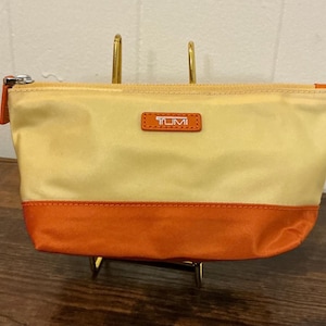 Peut inclure: Petite trousse de maquillage rectangulaire, partie supérieure dorée et partie inférieure orange. La trousse est dotée d'une fermeture éclair argentée et d'un petit patch en cuir avec la marque "TUMI". Présentée sur un support doré.
