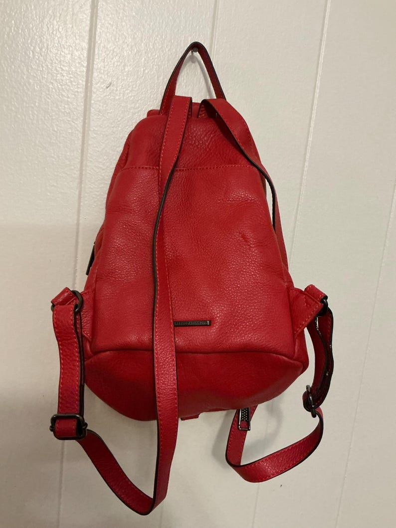 May include: A bright red backpack with a textured, pebbled leather-like finish. The bag features a top handle, adjustable shoulder straps with dark metal hardware, and a small rectangular metal plate centered on the front exterior pocket.