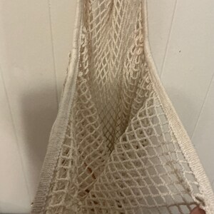May include: An off-white reusable market bag constructed from a woven cotton mesh material. The bag features a diamond-shaped net pattern throughout the body and includes a solid, reinforced fabric border along the top edge and the handle.