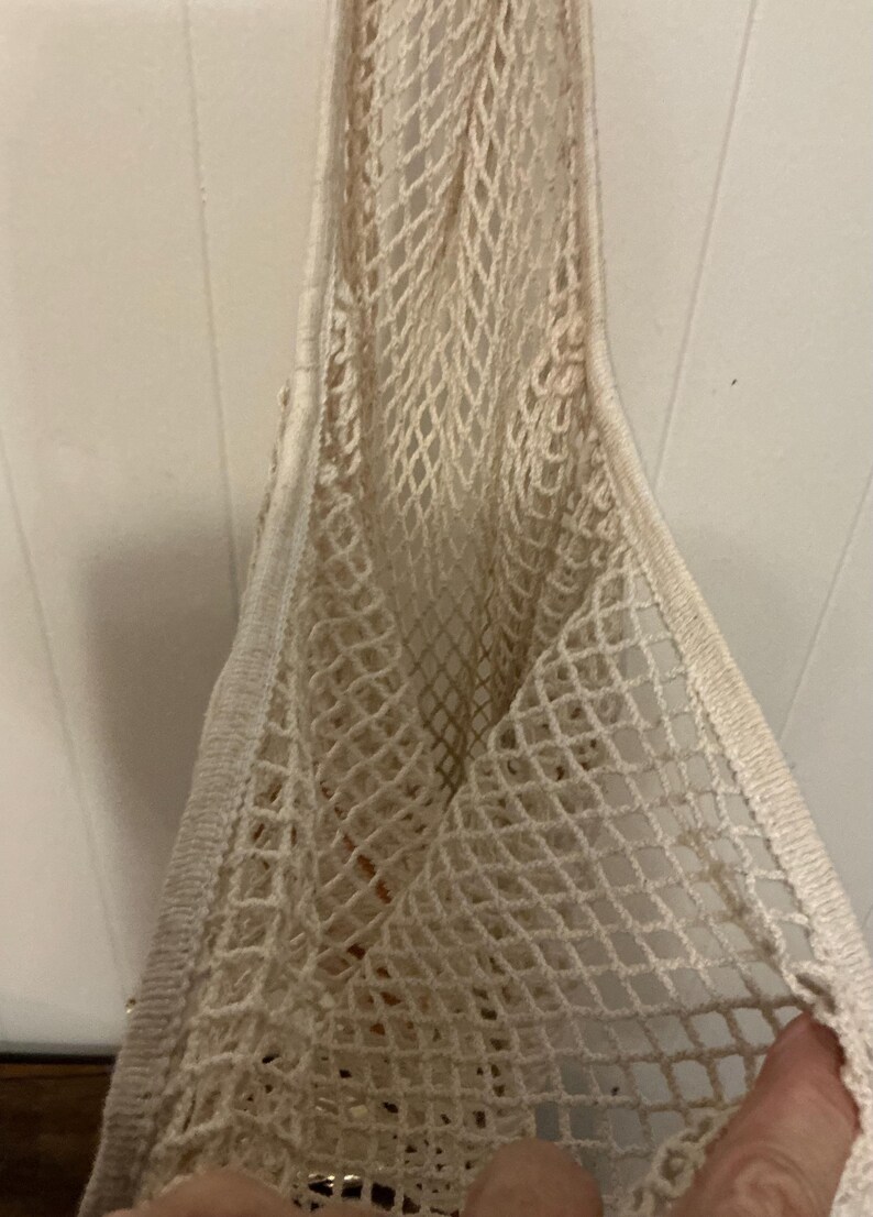 May include: An off-white reusable market bag constructed from a woven cotton mesh material. The bag features a diamond-shaped net pattern throughout the body and includes a solid, reinforced fabric border along the top edge and the handle.