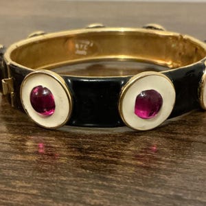 May include: A gold-toned hinged bangle bracelet with a black enamel band. The bracelet features white circles with a deep red gemstone in the center. The bracelet is on a wooden surface.