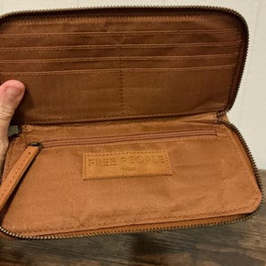 May include: Open brown leather wallet with multiple card slots and a zippered compartment. The wallet has a wrist strap and the brand name "FREE PEOPLE" is visible on a leather tag inside.