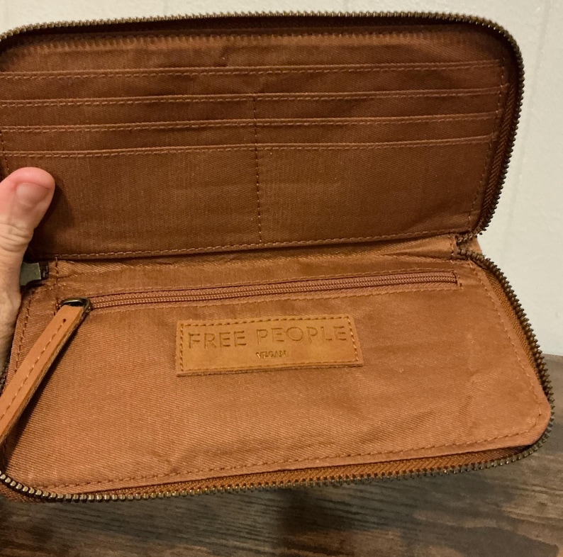 May include: Open brown leather wallet with multiple card slots and a zippered compartment. The wallet has a wrist strap and the brand name "FREE PEOPLE" is visible on a leather tag inside.