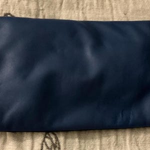 May include: A navy blue clutch purse with a rectangular shape. The purse is made of a smooth material and has a simple, minimalist design. The clutch is lying on a beige, patterned surface.