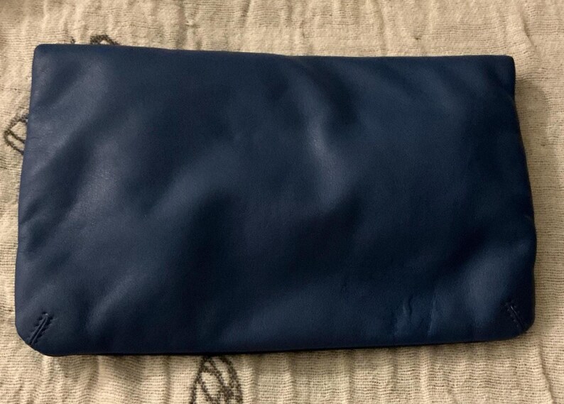 May include: A navy blue clutch purse with a rectangular shape. The purse is made of a smooth material and has a simple, minimalist design. The clutch is lying on a beige, patterned surface.