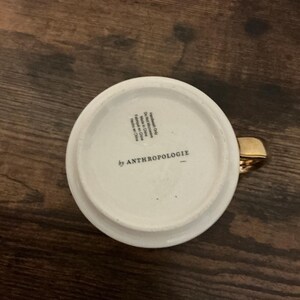 May include: White ceramic mug with a gold handle. The bottom of the mug features the text "by ANTHROPOLOGIE". The mug is sitting on a dark wooden surface.