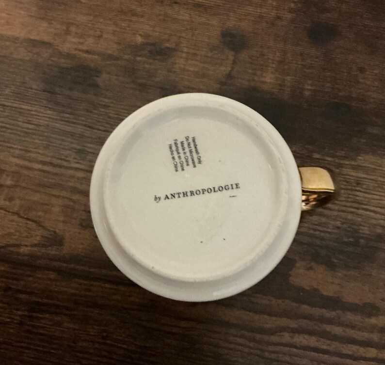 May include: White ceramic mug with a gold handle. The bottom of the mug features the text "by ANTHROPOLOGIE". The mug is sitting on a dark wooden surface.