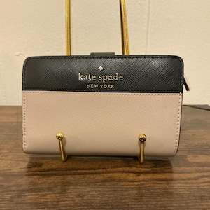 May include: A two-tone Kate Spade wallet in black and pale pink. The top section is black, and the bottom is pale pink. The brand name is in gold lettering. The wallet is displayed on a gold stand.