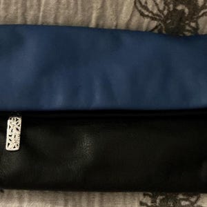 May include: A two-tone clutch with a blue top and black bottom. The clutch has a silver floral clasp. The bag is made of a soft, possibly leather, material. The clutch is folded over.