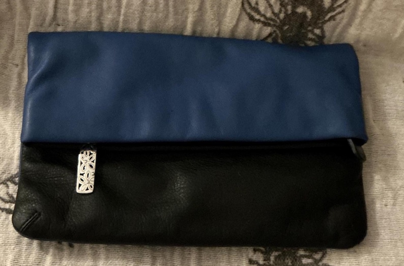 May include: A two-tone clutch with a blue top and black bottom. The clutch has a silver floral clasp. The bag is made of a soft, possibly leather, material. The clutch is folded over.
