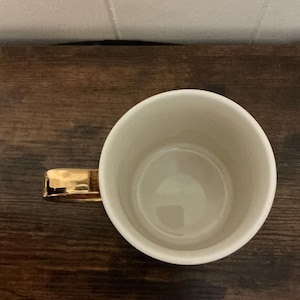 May include: A white ceramic mug with a gold-colored handle. The mug is empty and sits on a dark wooden surface. The handle is rectangular and appears to be made of metal. The interior of the mug is a light cream color.