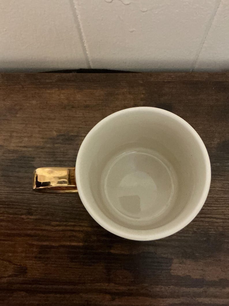 May include: A white ceramic mug with a gold-colored handle. The mug is empty and sits on a dark wooden surface. The handle is rectangular and appears to be made of metal. The interior of the mug is a light cream color.