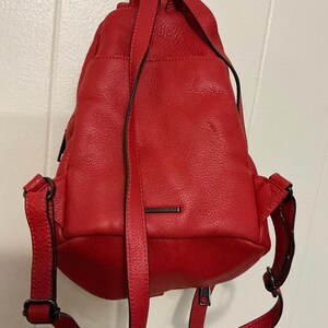 May include: A bright red leather backpack with a pebbled texture, featuring adjustable shoulder straps, a top handle, and a small rectangular metal plate with the brand name Rebecca Minkoff centered on the front exterior pocket.