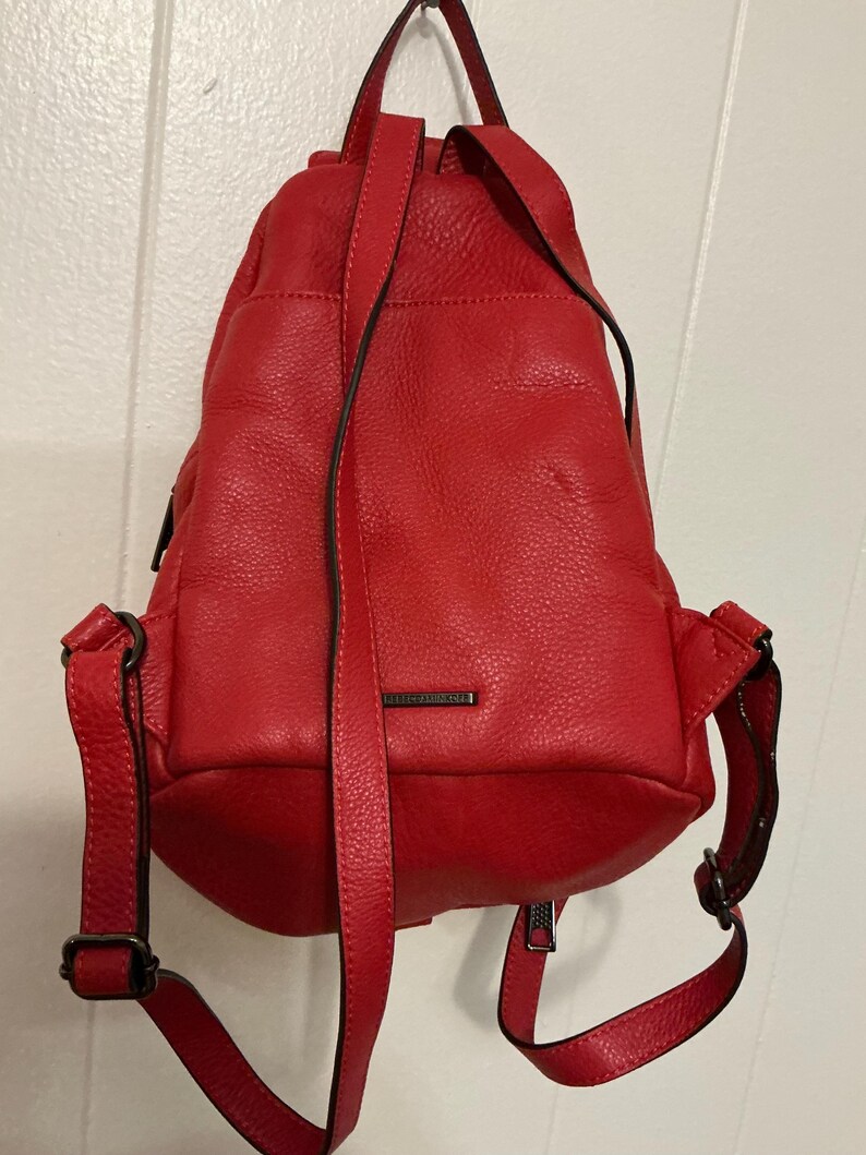May include: A bright red leather backpack with a pebbled texture, featuring adjustable shoulder straps, a top handle, and a small rectangular metal plate with the brand name Rebecca Minkoff centered on the front exterior pocket.