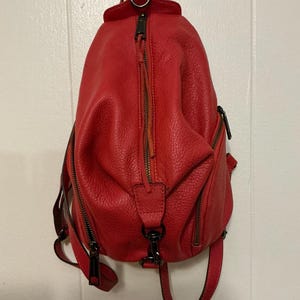 May include: A bright red textured leather backpack featuring a vertical zippered front pocket, two side zippered compartments, and dark metal hardware. The bag includes a top handle, adjustable shoulder straps, and a central metal clip detail.