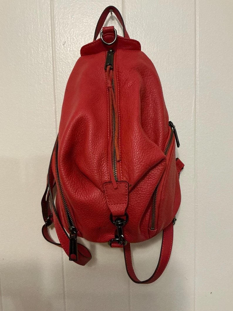 May include: A bright red textured leather backpack featuring a vertical zippered front pocket, two side zippered compartments, and dark metal hardware. The bag includes a top handle, adjustable shoulder straps, and a central metal clip detail.