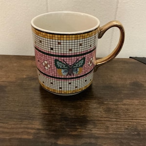 May include: A ceramic mug with a gold handle and rim. The mug features a pink and white checkered pattern with a butterfly design in the center. The mug is sitting on a wooden surface.