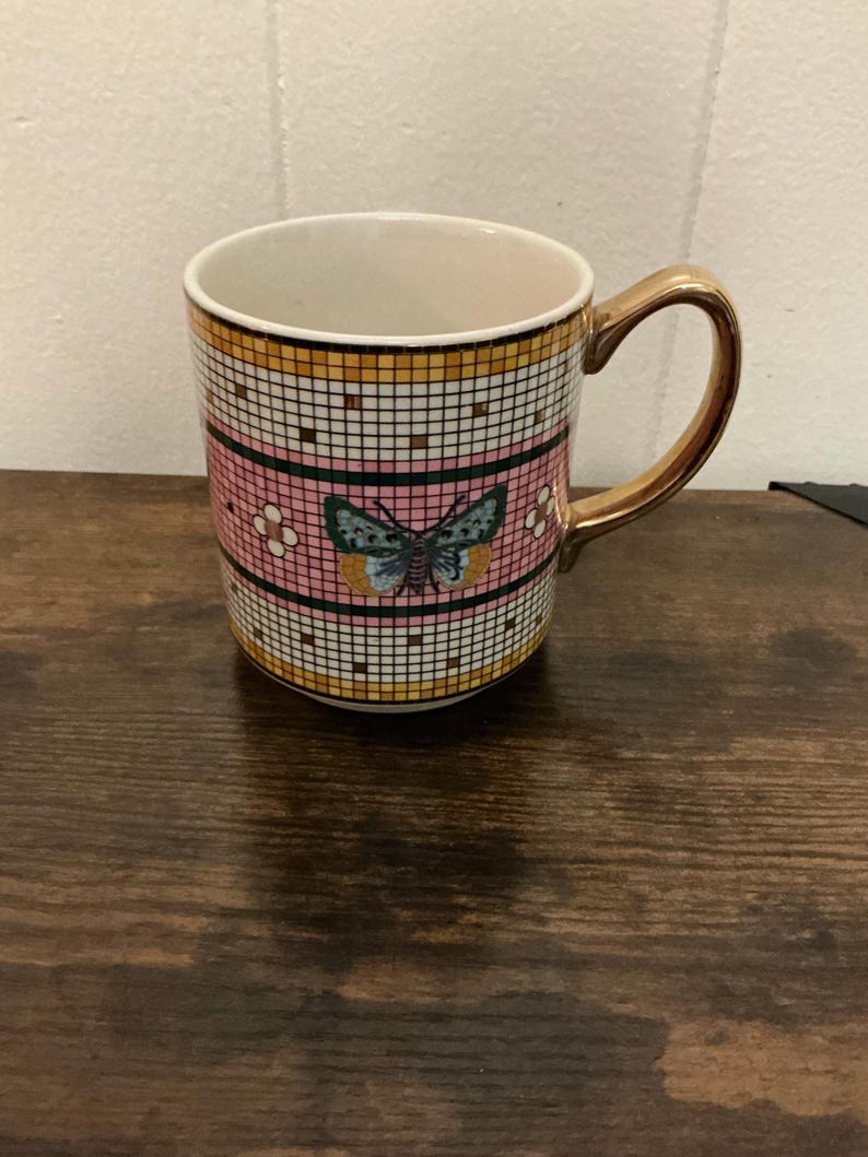 May include: A ceramic mug with a gold handle and rim. The mug features a pink and white checkered pattern with a butterfly design in the center. The mug is sitting on a wooden surface.