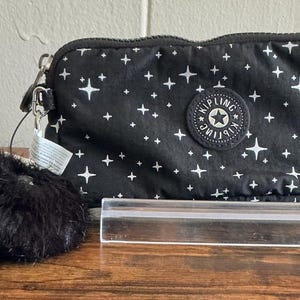 Peut inclure: Pochette noire zippée avec un motif d'étoiles blanches et un pompon noir duveteux. La pochette a une dragonne et un logo circulaire avec une étoile. Une étiquette avec le nom de la marque est attachée.