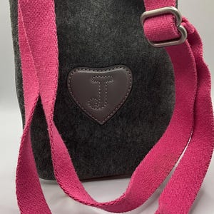 May include: Dark gray crossbody bag with a bright pink strap. The bag features a heart-shaped patch with the letter "J" on the front. The strap is adjustable and made of a sturdy fabric.