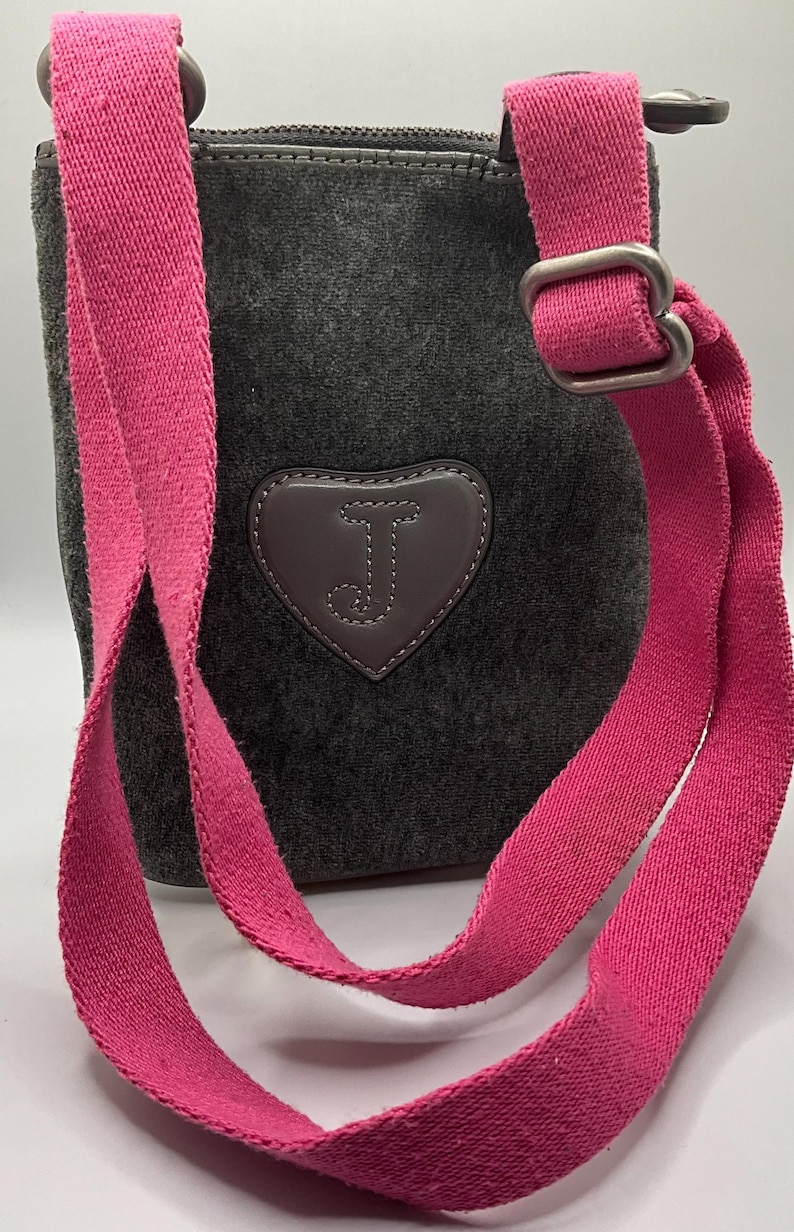 May include: Dark gray crossbody bag with a bright pink strap. The bag features a heart-shaped patch with the letter "J" on the front. The strap is adjustable and made of a sturdy fabric.