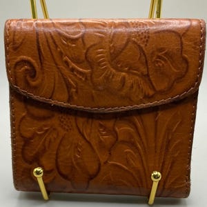 May include: A brown leather wallet with a floral embossed design. The wallet has a flap closure and is held by a gold-colored stand. The leather has a rich, warm tone, and the embossed pattern adds texture and visual interest.