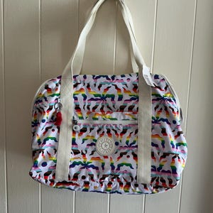 May include: A white tote bag with a colorful rainbow pattern. The bag has a zippered main compartment, a front pocket, and white handles. A small red tassel hangs from the zipper. The bag is hanging against a white wall.