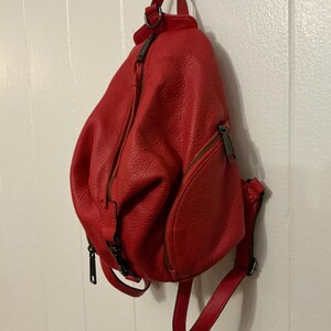 May include: A bright red, textured leather backpack featuring a teardrop shape, a vertical zippered main compartment, and a smaller front zippered pocket. The bag includes adjustable shoulder straps and dark metal hardware accents throughout.