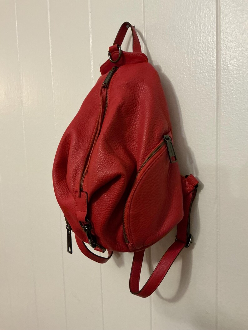 May include: A bright red, textured leather backpack featuring a teardrop shape, a vertical zippered main compartment, and a smaller front zippered pocket. The bag includes adjustable shoulder straps and dark metal hardware accents throughout.