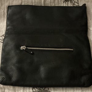 May include: Black leather clutch with a silver zipper closure. The clutch is rectangular and has a folded-over top. The leather has a textured appearance.