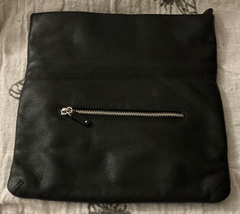May include: Black leather clutch with a silver zipper closure. The clutch is rectangular and has a folded-over top. The leather has a textured appearance.