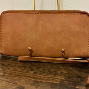 May include: A brown leather wallet with a full zipper closure and a wrist strap. The wallet has a rectangular shape and a smooth texture. Two gold-colored metal rings are attached to the front of the wallet.