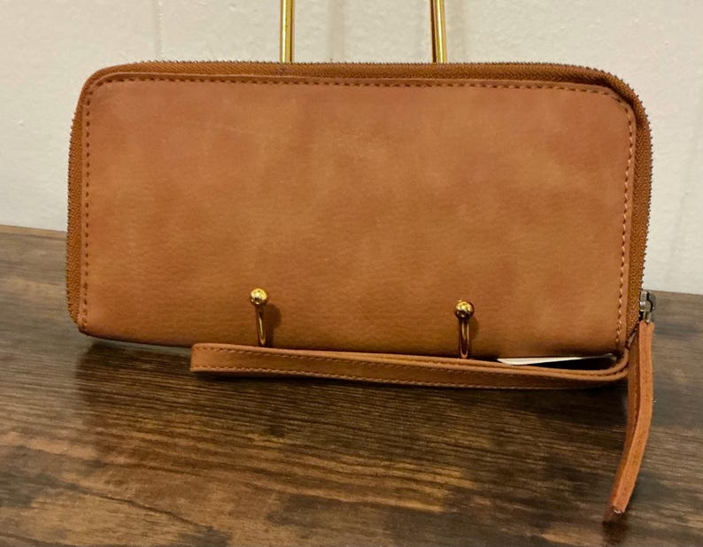 May include: A brown leather wallet with a full zipper closure and a wrist strap. The wallet has a rectangular shape and a smooth texture. Two gold-colored metal rings are attached to the front of the wallet.
