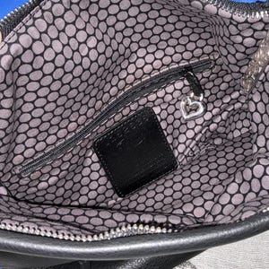 May include: Open view of a black handbag interior with a blue accent. The interior features a gray and black polka dot pattern lining. A small zippered pocket and a black leather tag are visible. A silver heart-shaped charm hangs from the zipper.