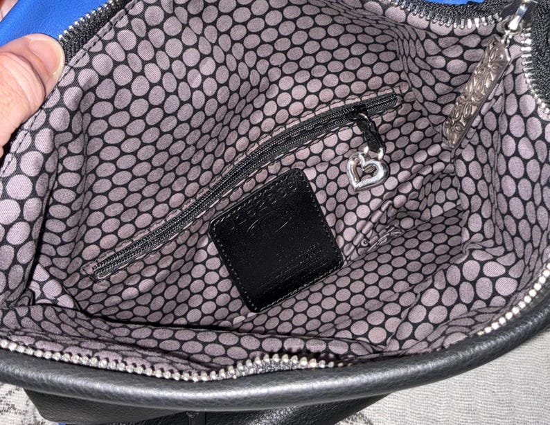 May include: Open view of a black handbag interior with a blue accent. The interior features a gray and black polka dot pattern lining. A small zippered pocket and a black leather tag are visible. A silver heart-shaped charm hangs from the zipper.